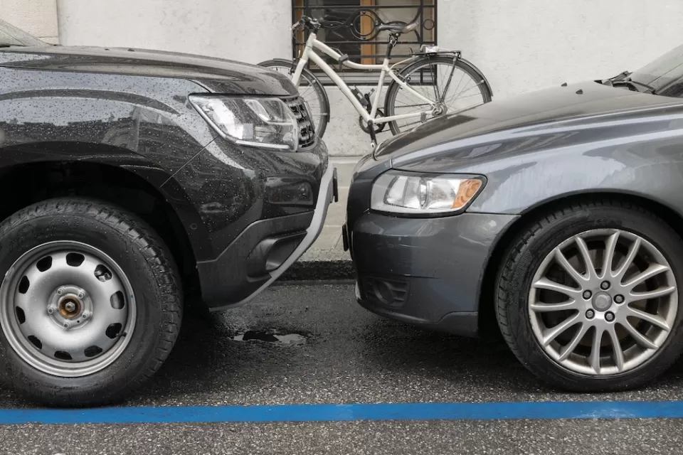 Two grey cars parked nose to nose, one taller than the other, on city curb with parked bicycle visible on sidewalk.